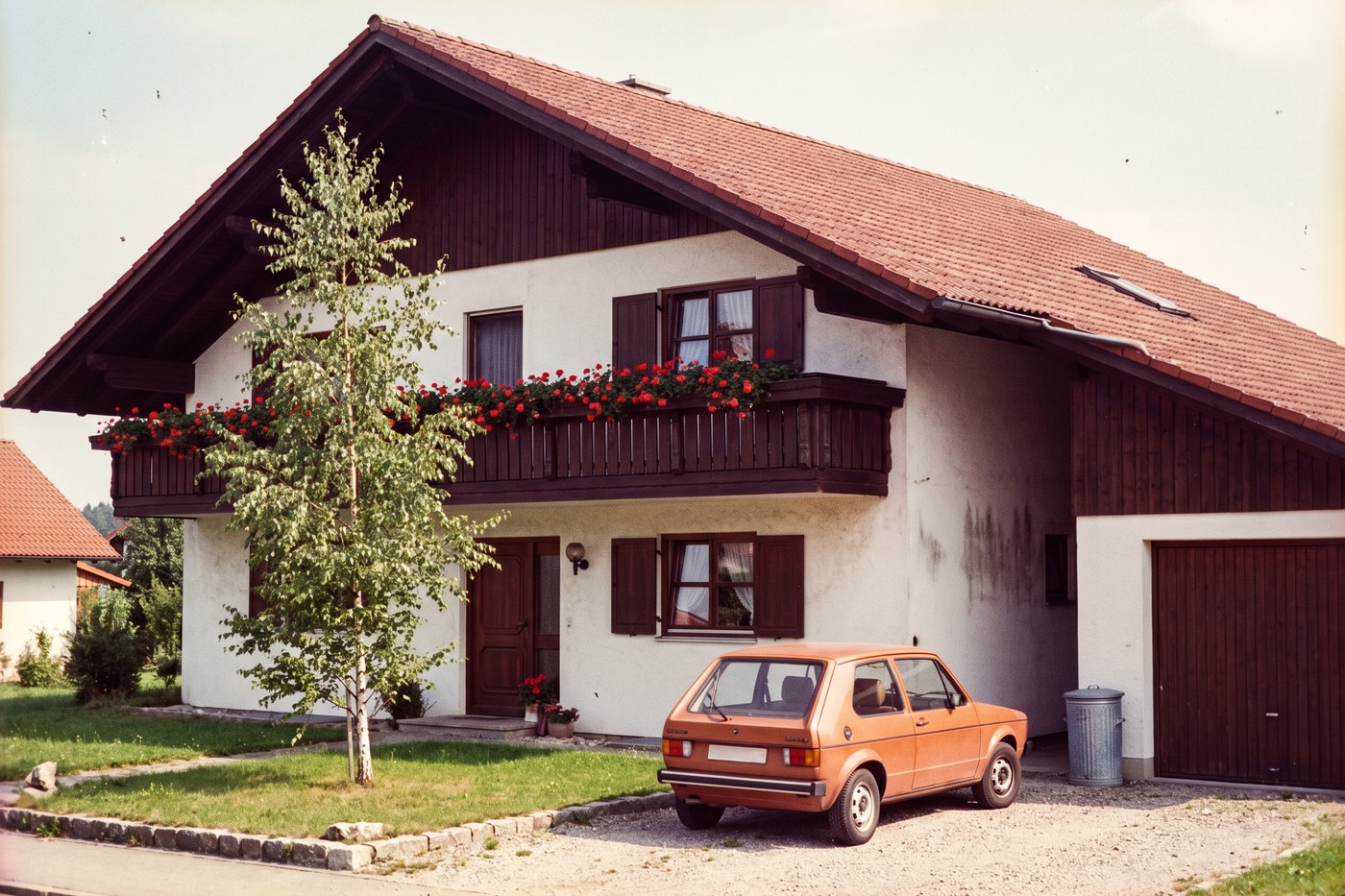 Altes, verblasstes Farbfoto aus einem Fotoalbum: Haus mit Holzbalkon und Geranien, davor ein oranges Auto und eine Mülltonne aus Blech