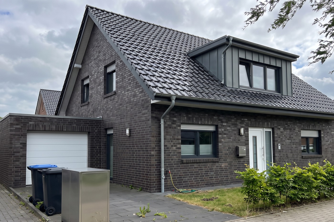 Foto eines neuen Hauses aus dunklem Klinker mit großer Gaube und Garage, davor Mülltonnen und eine Paketbox