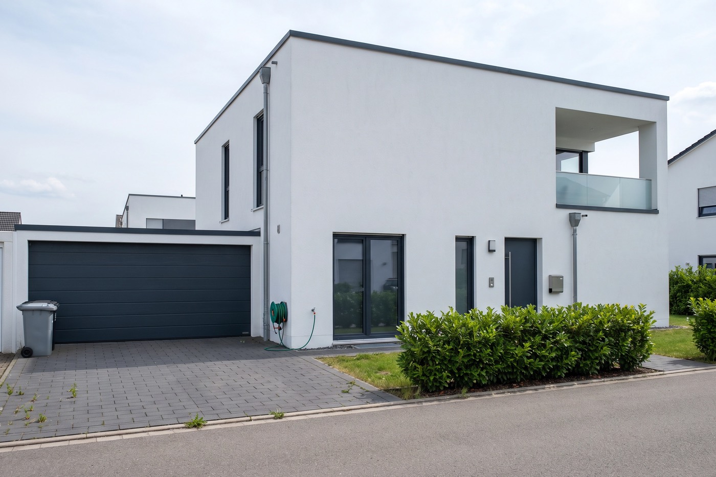Foto eines weißen Hauses im Bauhausstil mit Flachdach, Loggia und Doppelgarage, davor Mülltonnen und ein Gartenschlauch