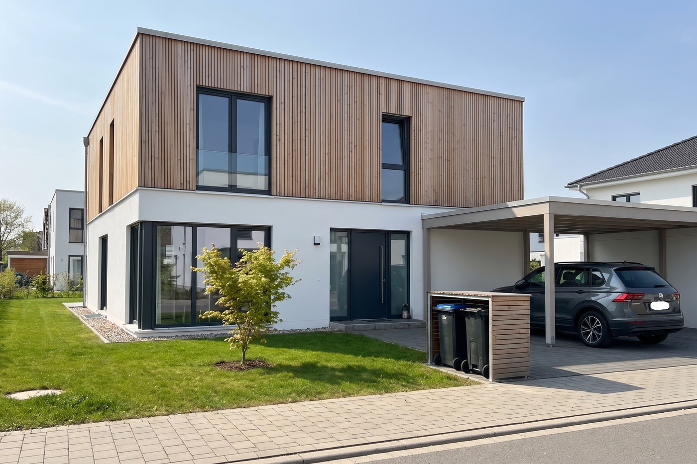 Foto eines modernen Neubaus mit Holzfassade und Carport, darunter ein Auto und eine Box mit Mülltonnen