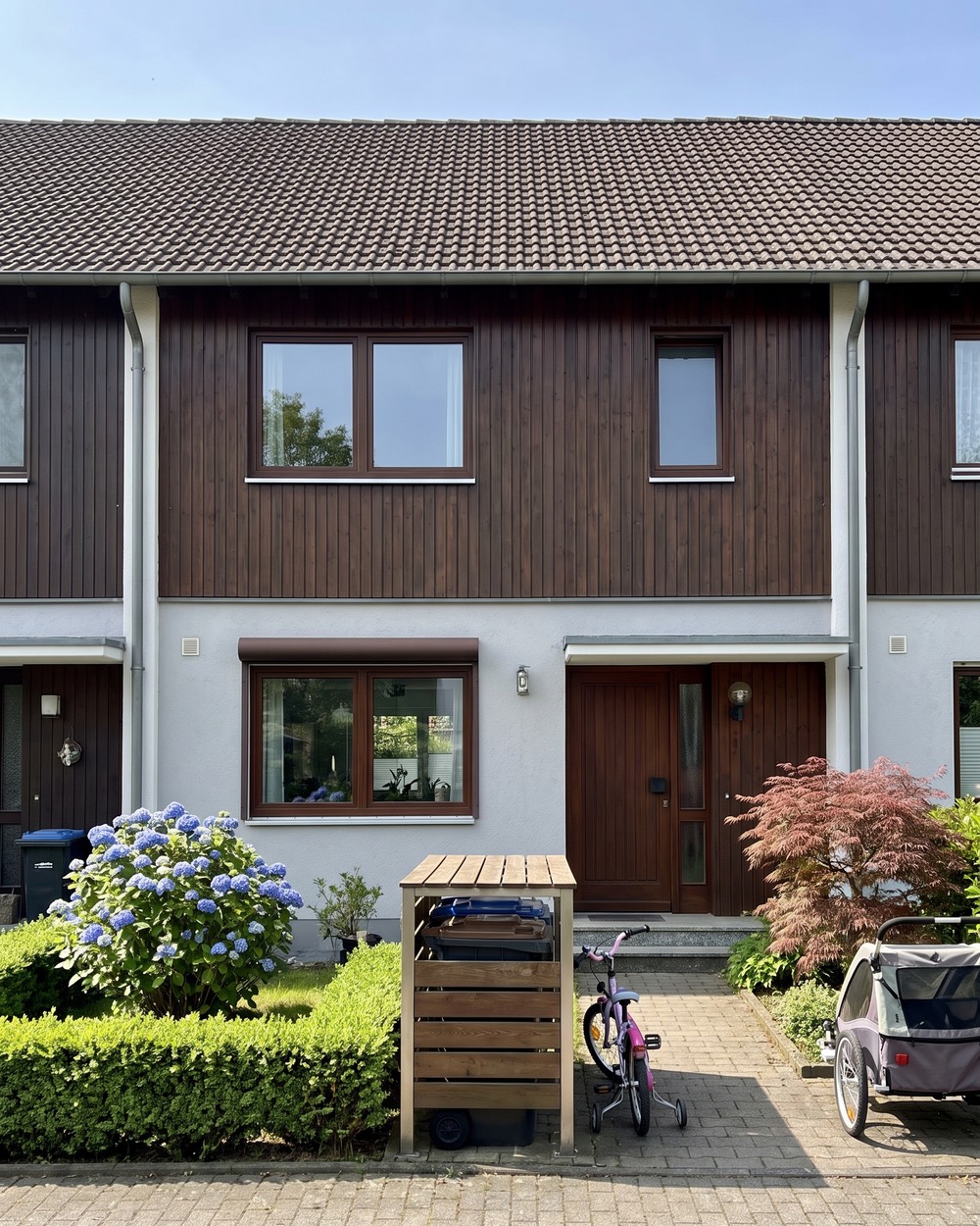 Foto eines Reihenmittelhauses mit dunkler Holzfassade oben, davor eine Box für Mülltonnen, ein Kinderfahrrad und ein Fahrradanhänger