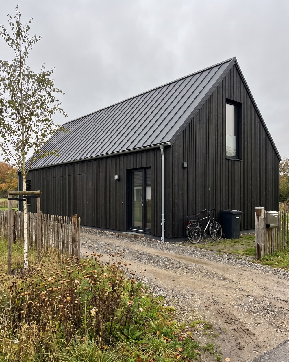 Foto eines Scheunenhauses mit schwarzer Holzfassade und Blechdach, an der Wand ein Fahrrad und eine Mülltonne
