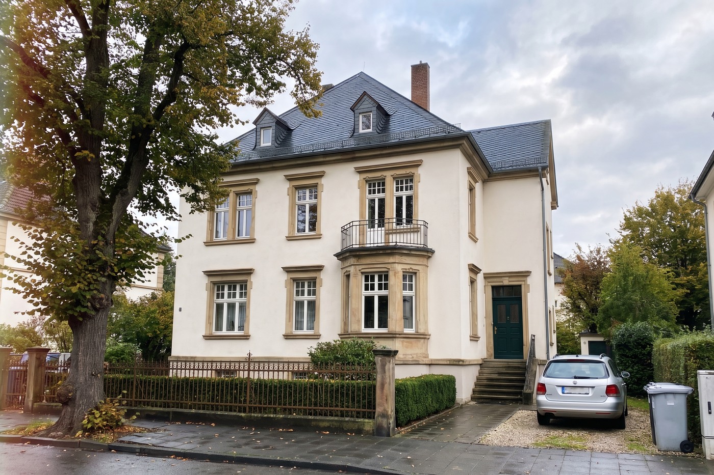 Foto einer weißen Stadtvilla mit Schieferdach und Erker, davor ein silbernes Auto und eine Mülltonne